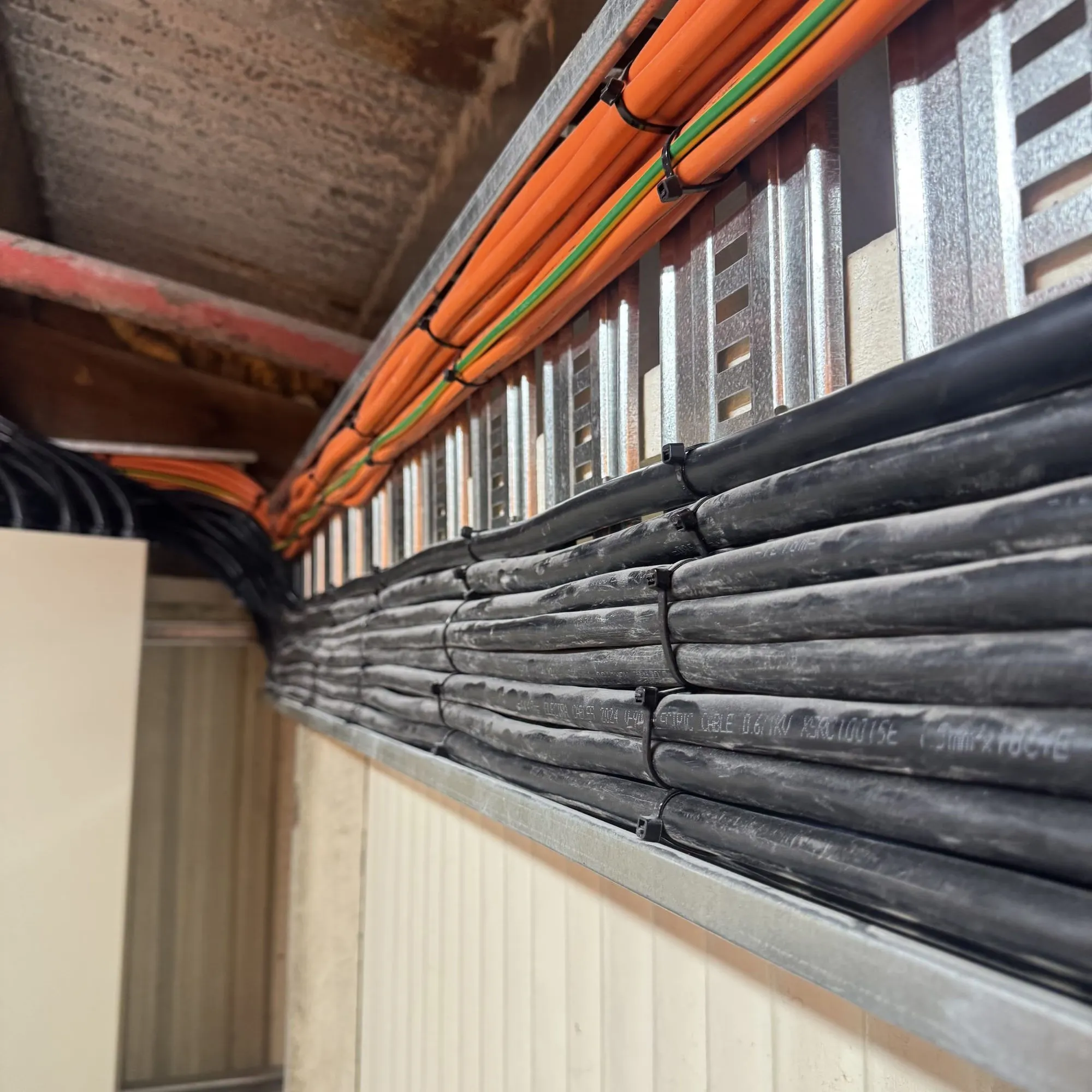 Black multicore cables and orange conductors cable-tied along a galvanised ladder rack on a wall