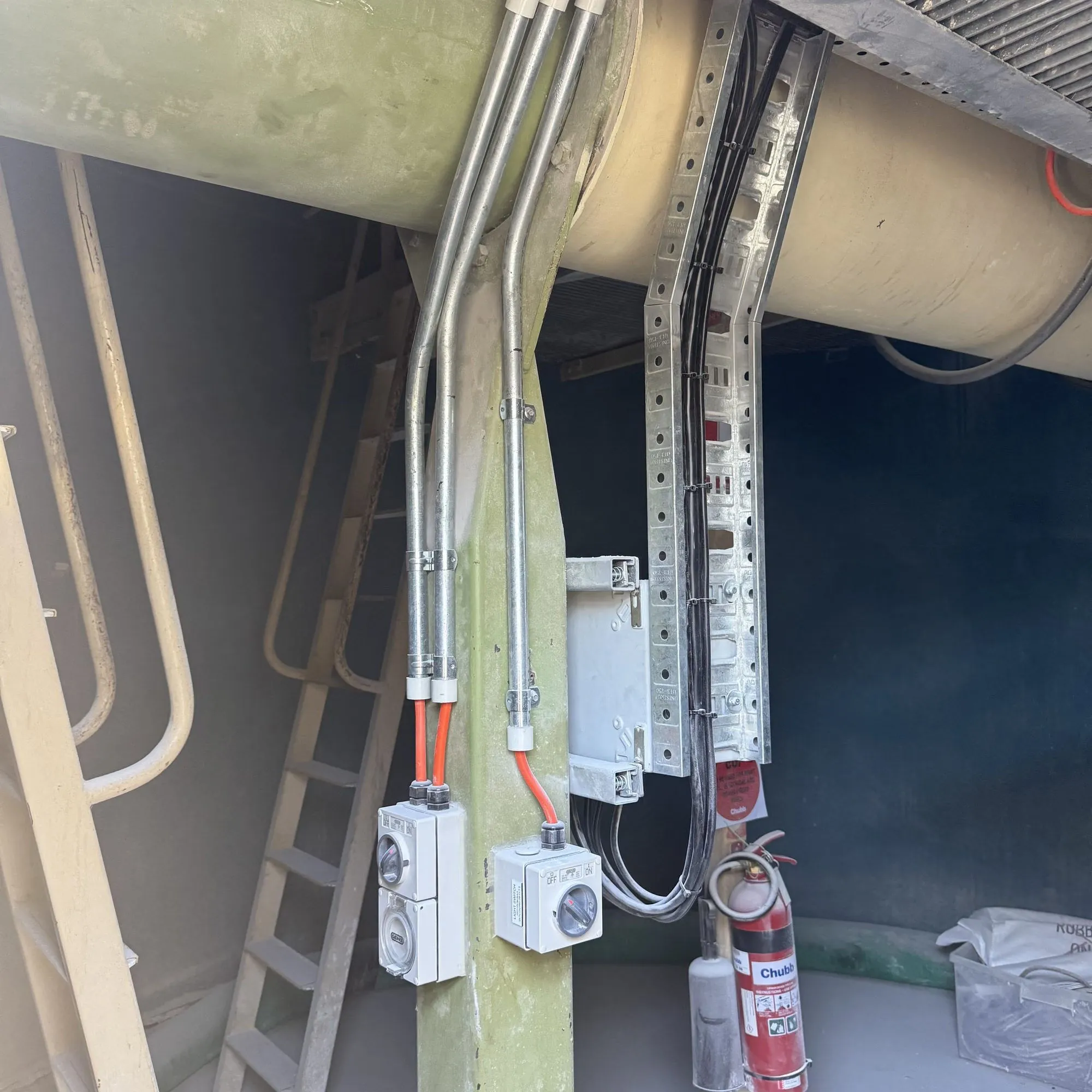 Galvanised conduit drops to weatherproof junction boxes on a silo support leg, beside a fire extinguisher