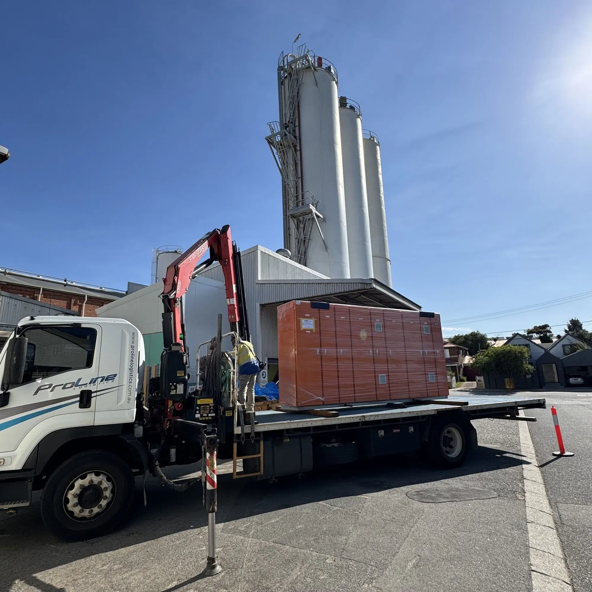 Crane truck delivering a crated orange switchboard on site, mill silos behind under clear sky
