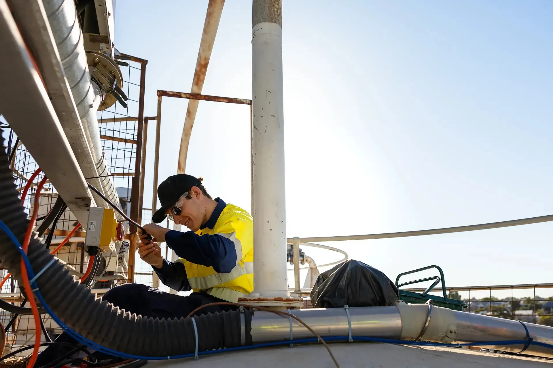 Electrician crouched beside flexible conduit and ductwork, working on cabling atop an outdoor plant structure