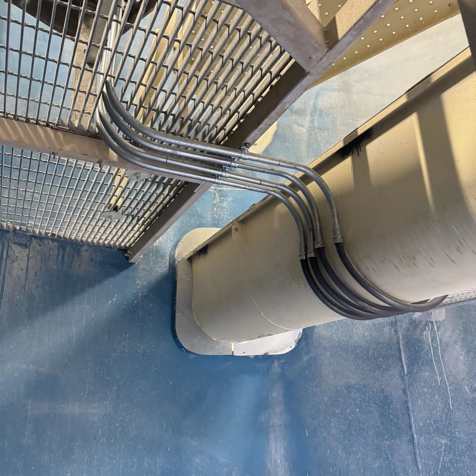 Four galvanised conduits swept in parallel bends beneath a steel grated walkway on plant structure