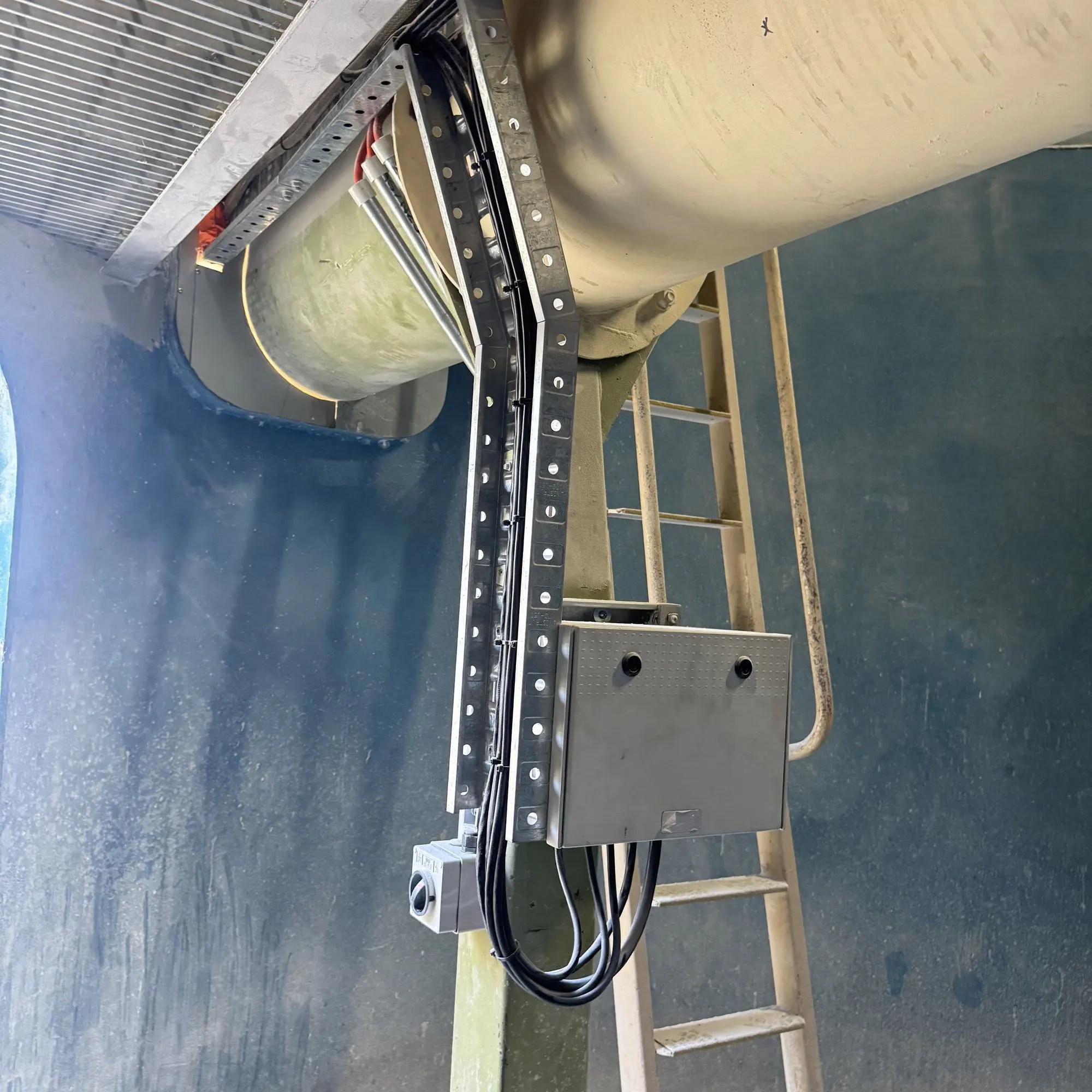 Junction box and looped cables on a cable tray beside plant pipework, fixed ladder to one side