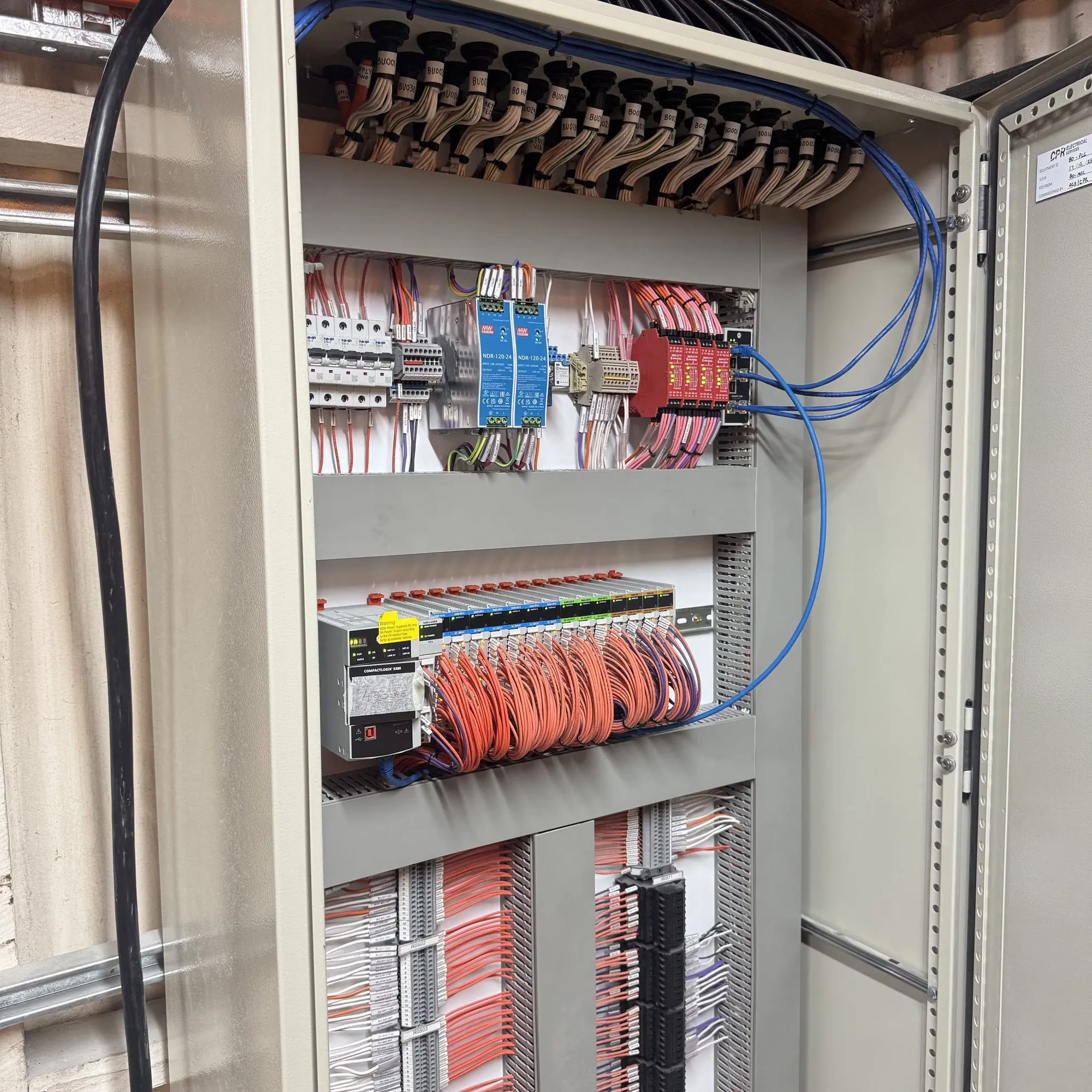 Open control cabinet showing breakers, power supplies, a PLC input rack and looped orange field wiring