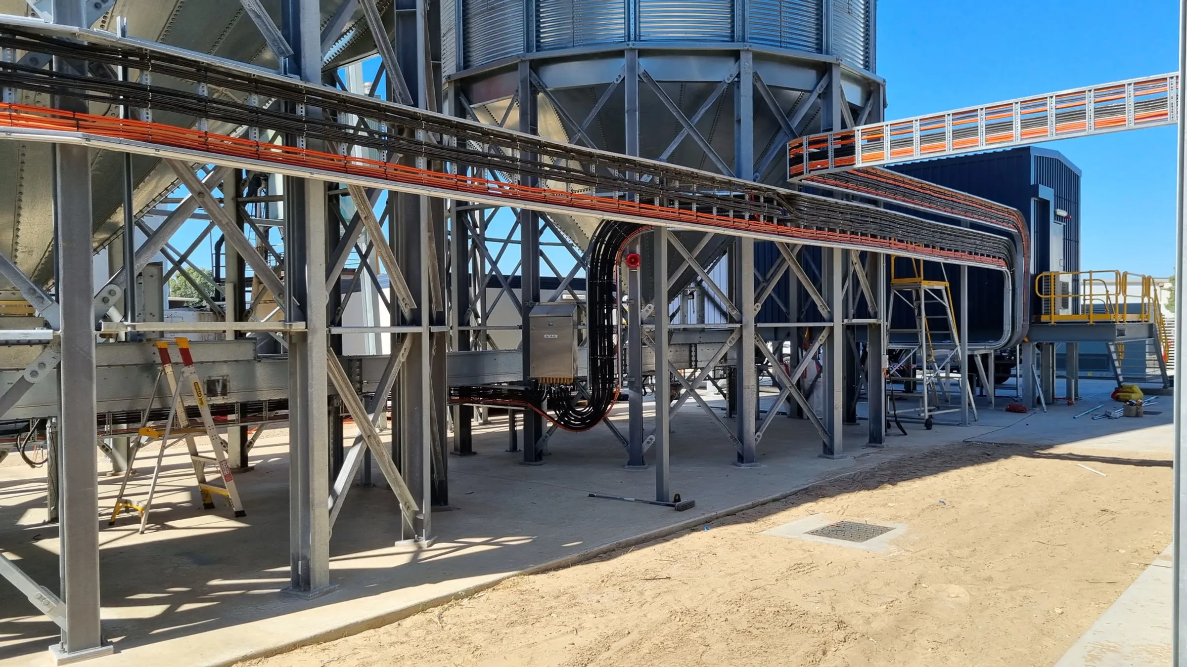 Orange cable ladder running along a steel gantry beneath grain silos at an outdoor processing plant